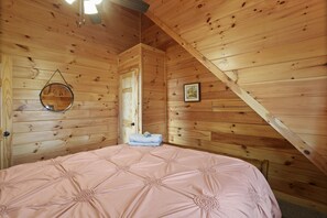 A bedroom with a wooden sloped ceiling, a bed with a pink bedspread, a circular mirror, and a framed picture on the wall.