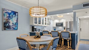 Un coin repas avec une table ronde et six chaises, un luminaire suspendu en rotin et une cuisine en arrière-plan.