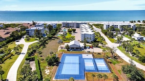 Vista aérea de una zona residencial costera con una gran cancha de tenis, varios edificios y una playa al fondo.