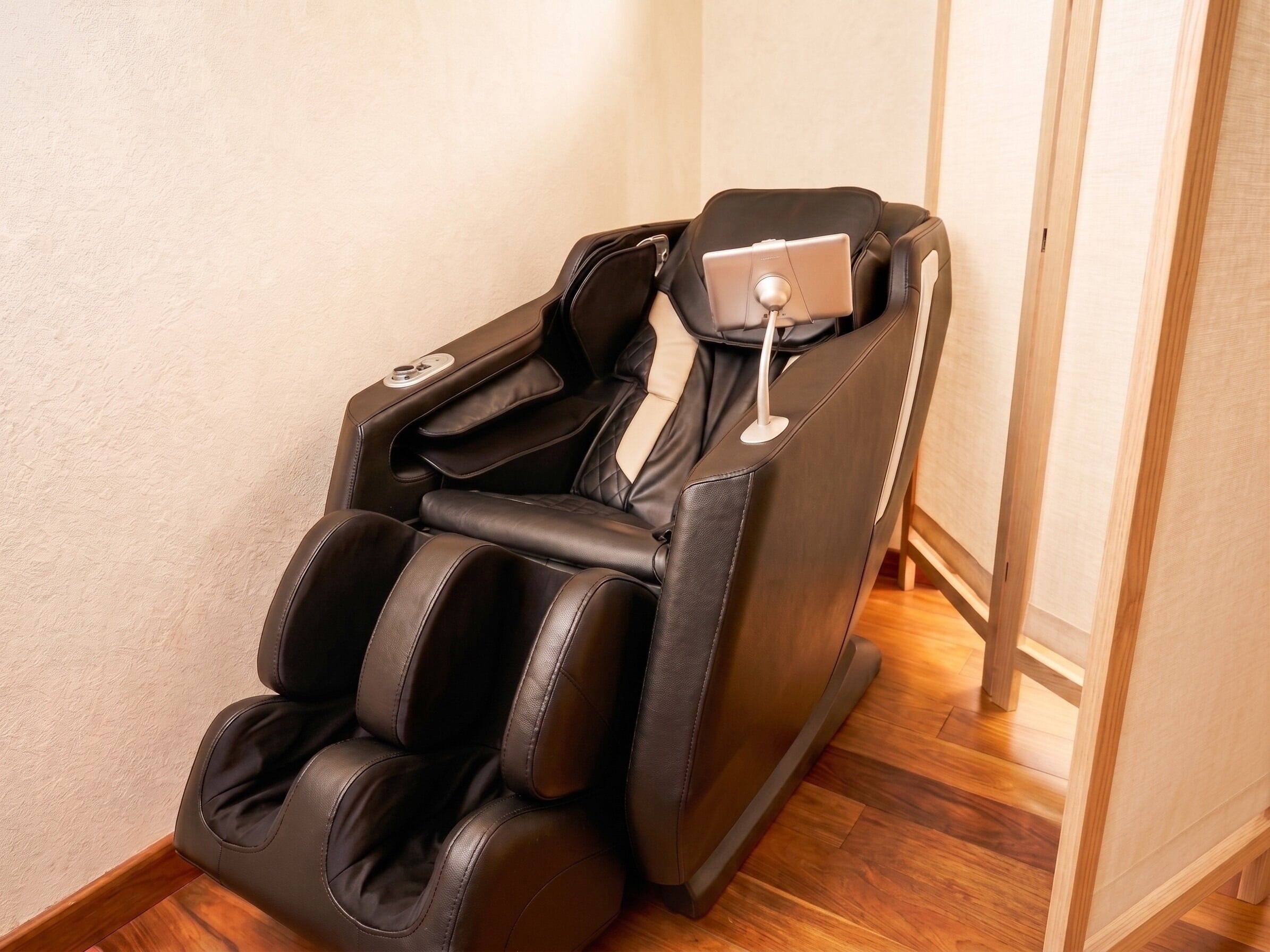 A massage chair with a control panel, positioned against a wall with wooden flooring.