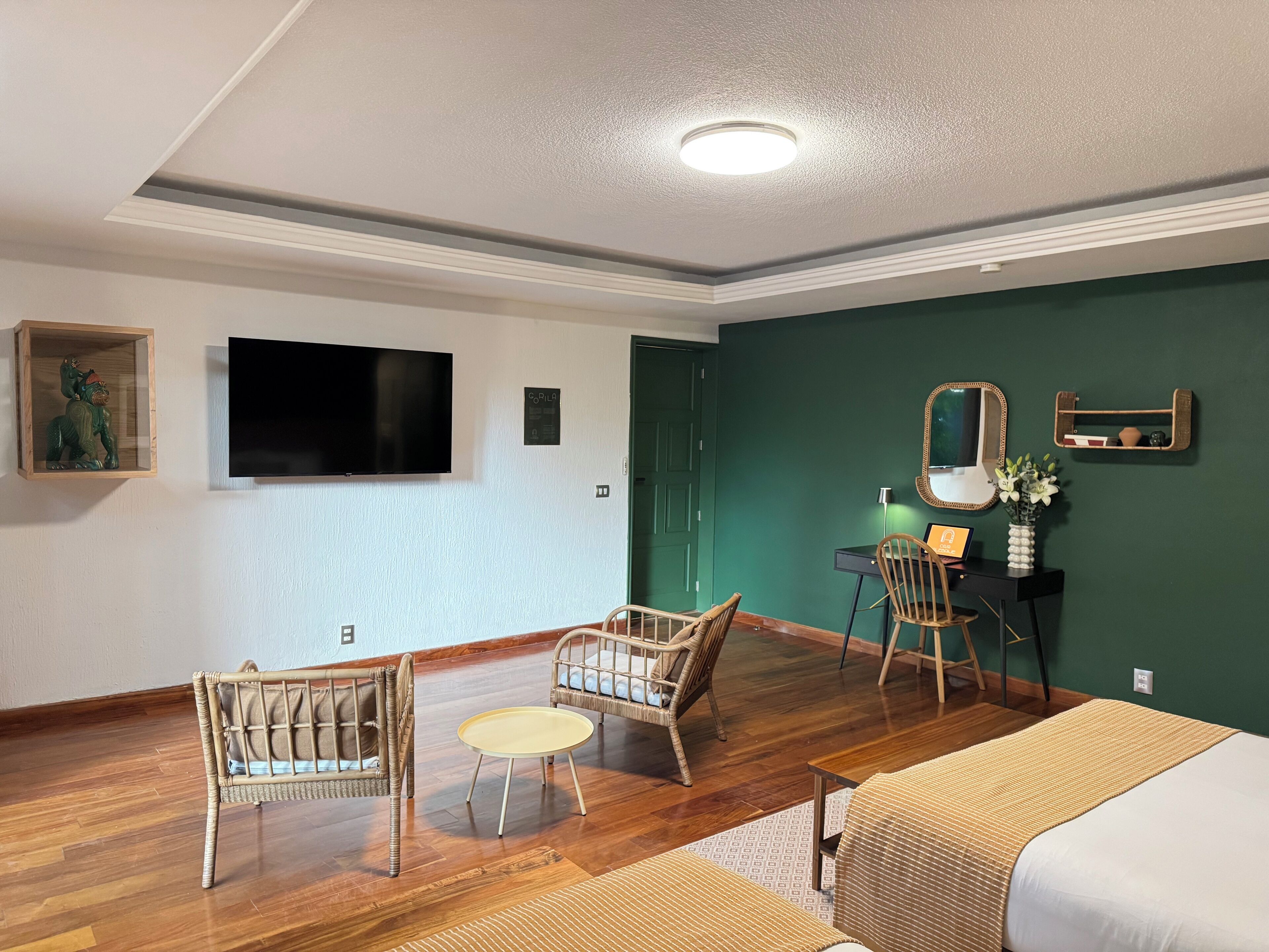 A room with a wooden floor, two wicker chairs, a small round table, a bed with a patterned bedspread, a desk with a chair, a flat-screen TV, and a green accent wall.