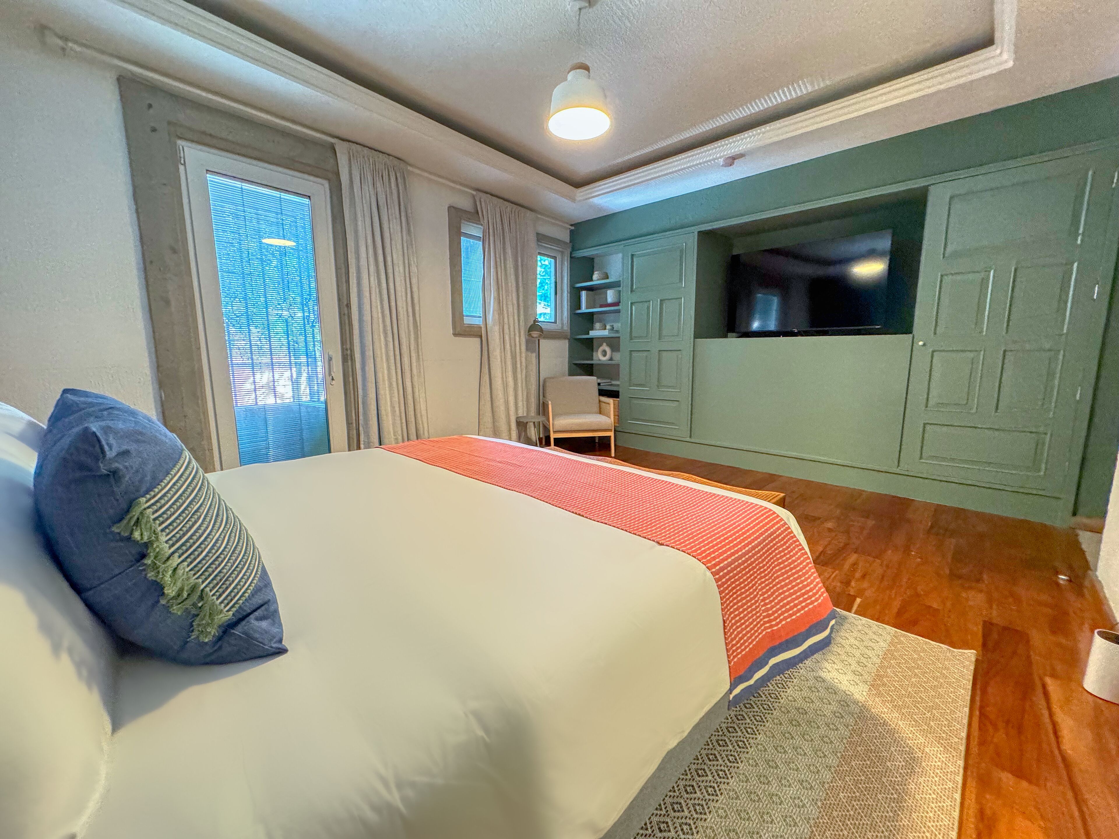Quarto com cama, cadeira, escrivaninha e parede verde com televisão integrada.