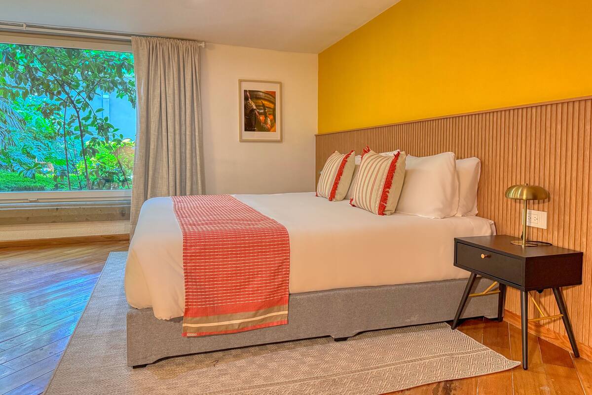 Une chambre d’hôtel moderne avec un grand lit, une table de chevet, une lampe, un tableau encadré au mur et une fenêtre donnant sur un paysage verdoyant.