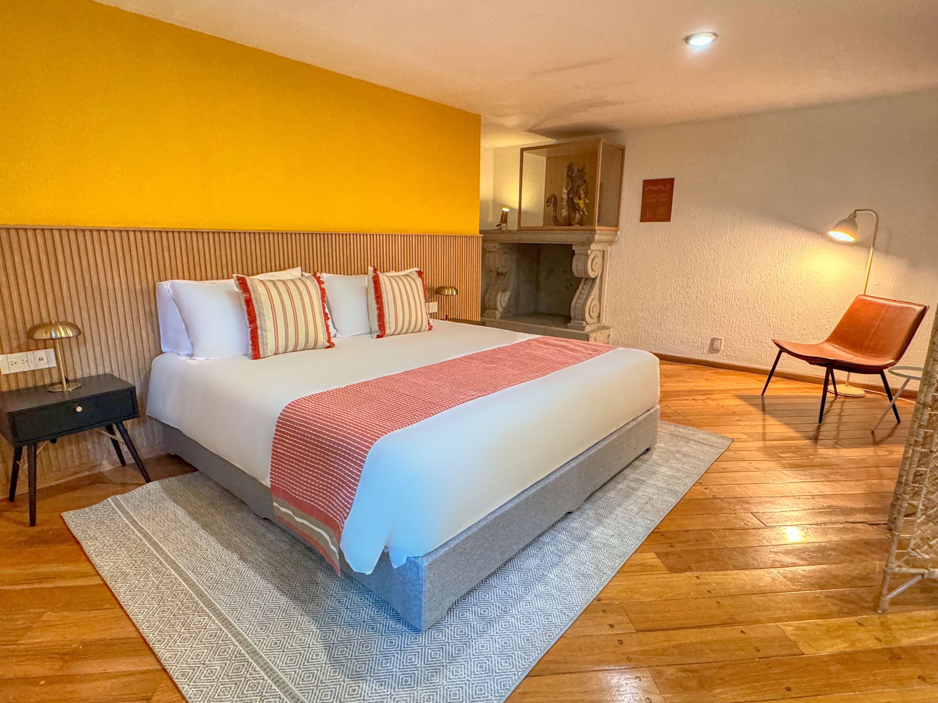 A hotel room with a large bed, a wooden floor, a yellow accent wall, a small table with a lamp, and a chair.