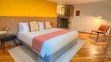 A hotel room with a large bed, a wooden floor, a yellow accent wall, a small table with a lamp, and a chair.