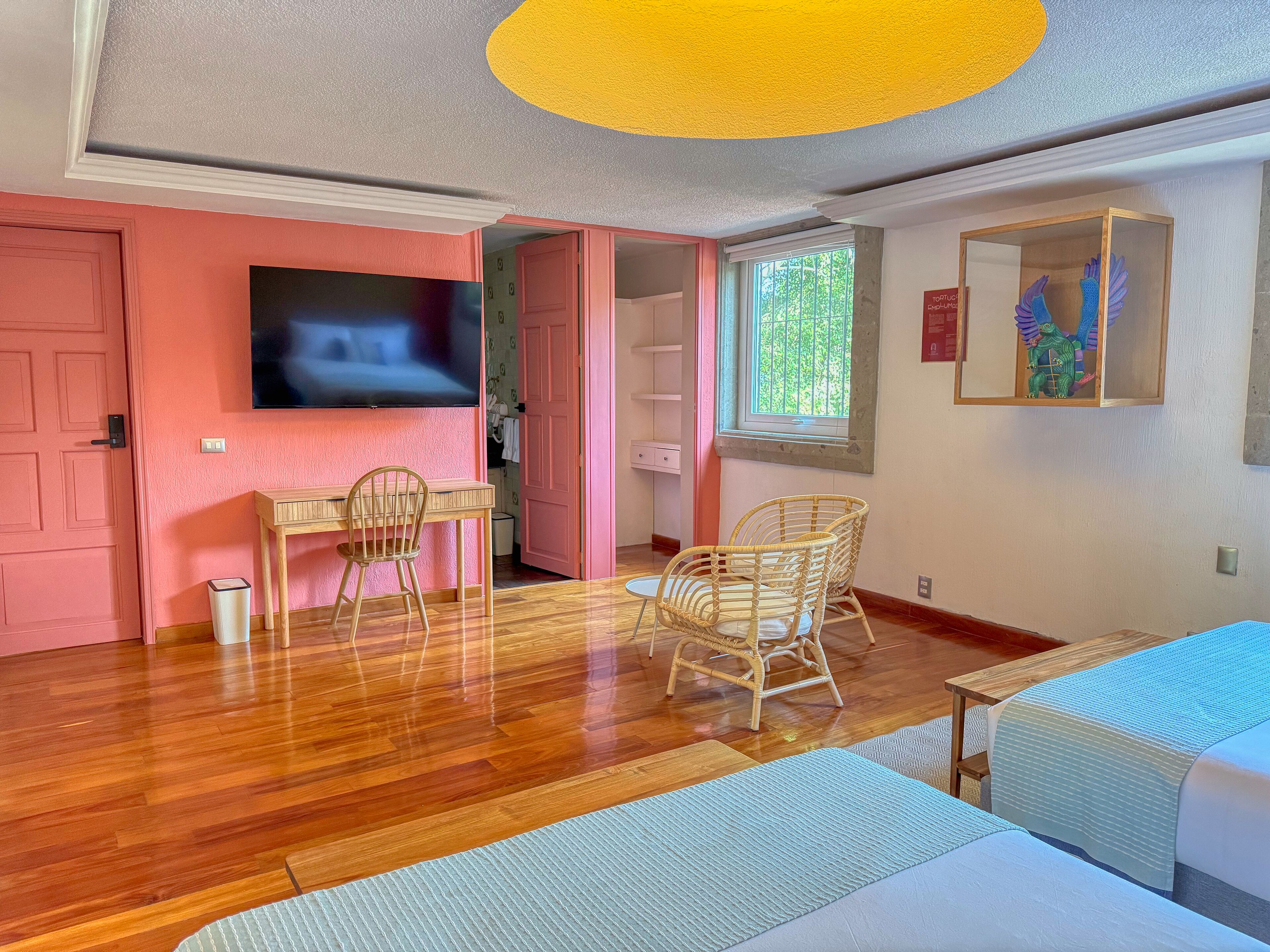 Una stanza con pavimento in legno, una TV montata a parete, una scrivania con una sedia, due poltrone in vimini e un letto con copriletto blu.