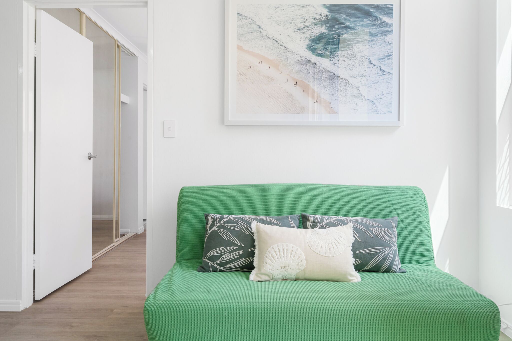 A green sofa with patterned pillows, a framed artwork of a beach scene, and a wooden floor.