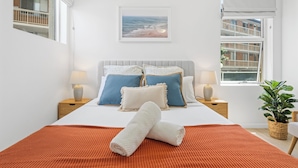 Een slaapkamer met een bed, kussens, een deken, een nachtkastje, een lamp, een raam en een plant.