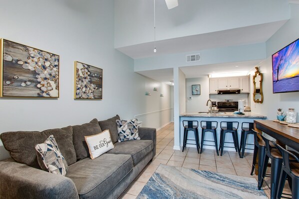 A modern living room with a grey sofa, a coffee table, and a kitchen area with a bar and stools. - Sugar Beach 368 Two-Story Beachside Retreat (Orange Beach)