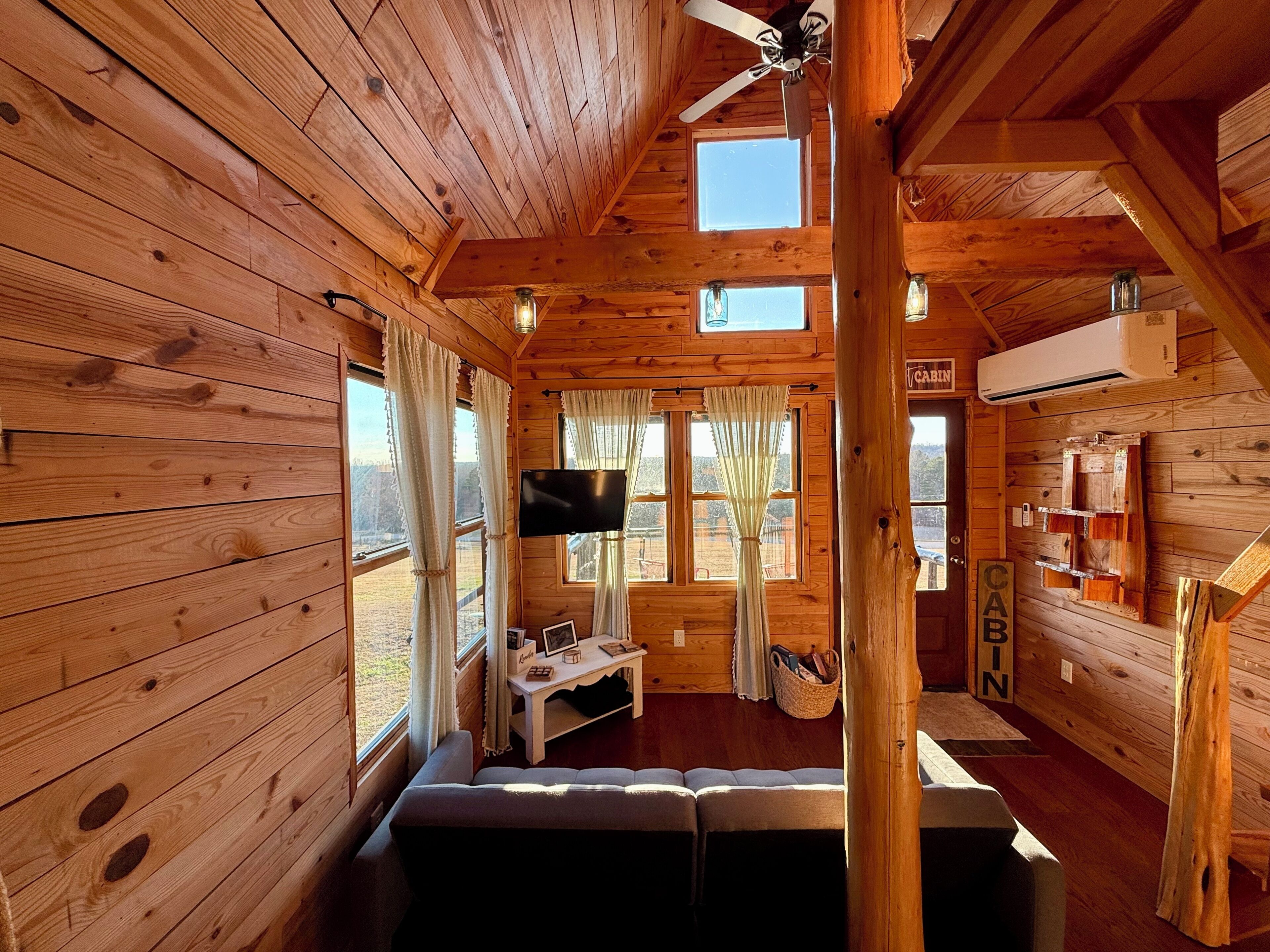 Une cabane aux murs en bois, avec un ventilateur de plafond, une télévision, un canapé et une fenêtre avec des rideaux.