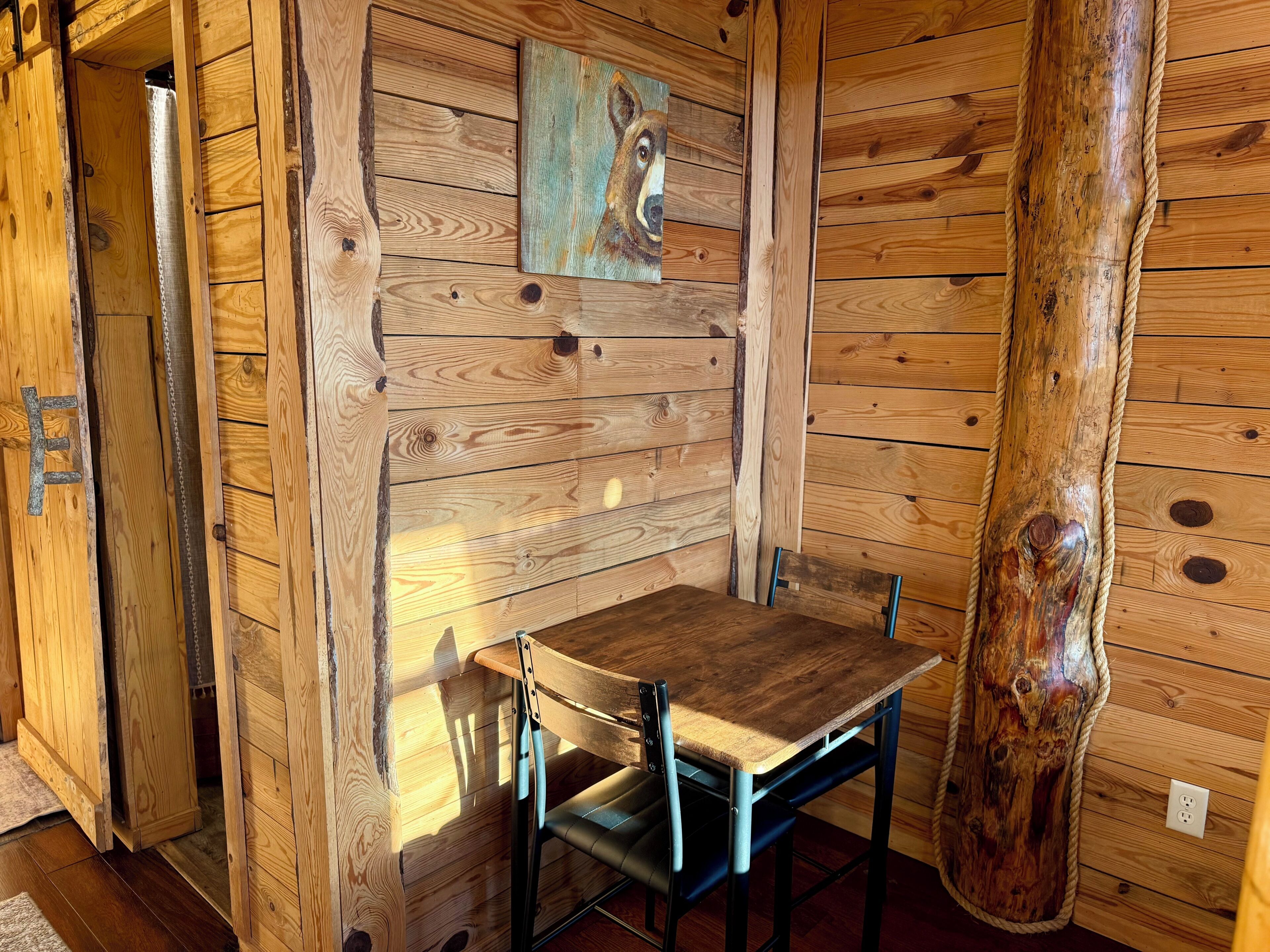 Ein rustikaler Holzraum mit Holztisch und Stuhl, einem Gemälde an der Wand und einem großen Baumstamm.