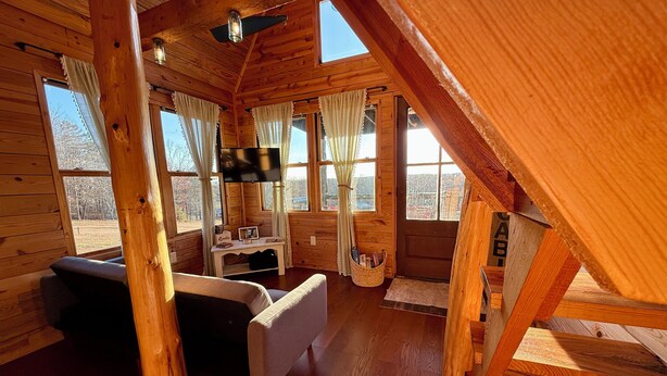 A wooden cabin interior with a sofa, a small table, a TV, and a window overlooking a snowy landscape.