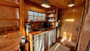A wooden cabin interior with a kitchen, wooden walls, a window, and a door.