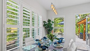 A dining area with a glass table, white chairs, and a view of greenery outside. - New Heated Pool Home in Fort Lauderdale,Charming Tropical Retreat Near Beaches, Shopping & Dining! (Fort Lauderdale)