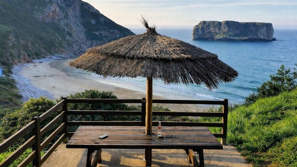 Un banc en bois, surmonté d’un parasol en chaume, offre une vue sur une plage bordée de falaises et sur une île lointaine.