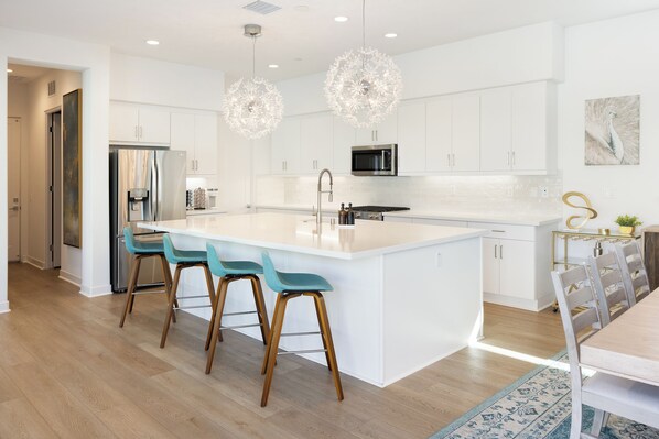 A modern kitchen with white cabinetry, a large island with bar stools, stainless steel appliances, and decorative crystal pendant lights. - Palm Oasis: Luxurious home in La Quinta with private heated pool and stunning mountain views. (La Quinta)