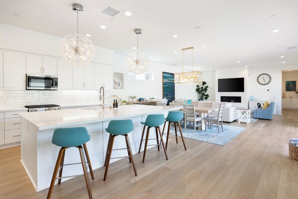 Cocina moderna con una isla central, taburetes de bar, zona de comedor y sala de estar con televisión y reloj.