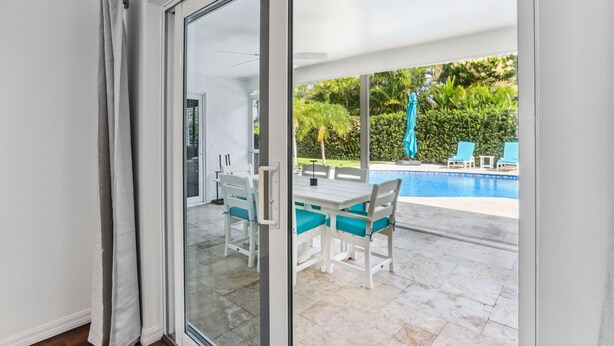 A room with a sliding glass door leading to an outdoor area with a pool, a table with chairs, and a view of greenery.