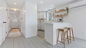Une cuisine moderne avec des meubles blancs, un îlot central, des tabourets en bois et une fenêtre qui laisse entrer la lumière naturelle.