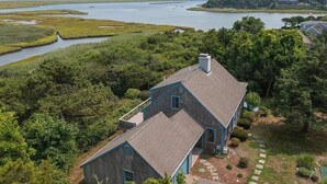 Une maison avec un toit à pignon, une cheminée et un porche à l’avant. Elle est entourée d’une végétation luxuriante et bordée par un plan d’eau.