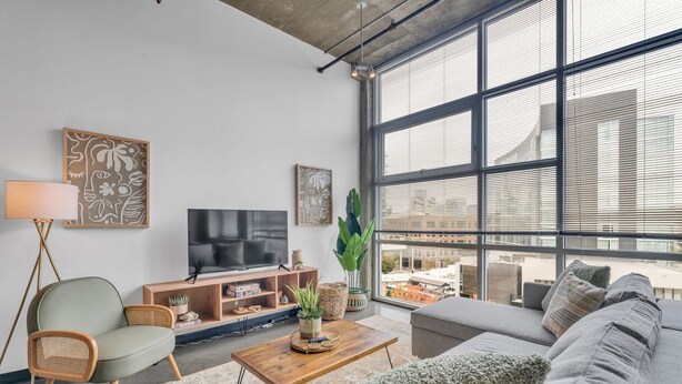 A modern living room with a large window, a flat-screen TV, a sofa, a coffee table, and decorative items. - Downtown 1 Bedroom Loft in the Gulch (Nashville)