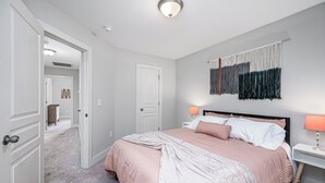 A bedroom with a large bed, two bedside lamps, a wall-mounted tapestry, and a door leading to another room. - Adorable home in fantastic location near restaurants and Duke! (Durham)