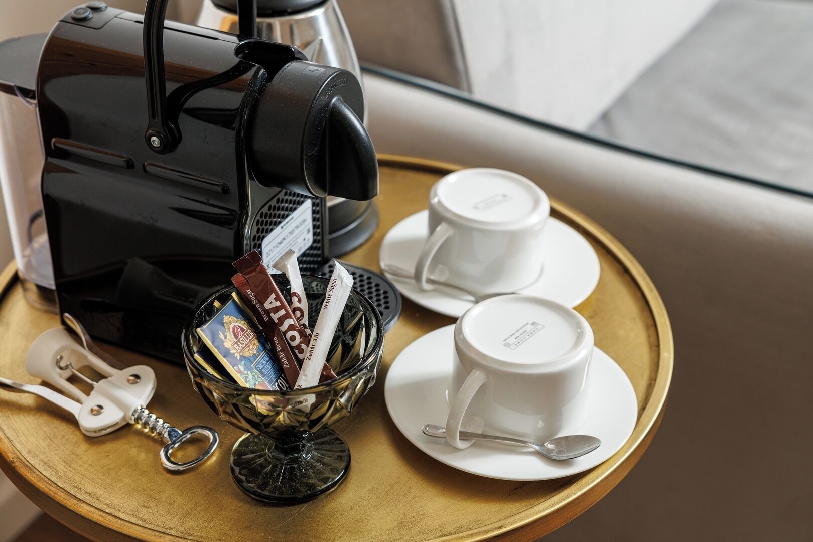 A coffee machine, two white cups, a glass bowl with coffee pods, and a spoon on a tray.