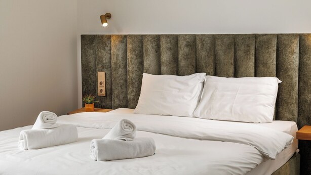 A neatly made bed with white linens and two rolled towels, a wooden headboard, and a small bedside table with a plant. - Richter Residence - Apartment 8 (Brașov)