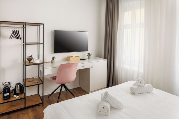 Quarto de hotel com uma cama, uma secretária com televisão, uma cadeira rosa, um guarda-roupa e uma janela com cortinas.