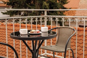 Uma mesa redonda com uma cadeira de vime, um tabuleiro com macarons e duas chávenas brancas, e um guarda-corpo de varanda com telhado de azulejos.