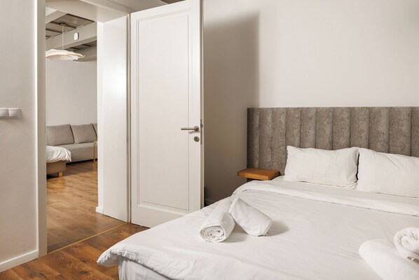 Una habitación de hotel con una cama, una mesita de noche, suelo de madera, un cabecero tapizado en gris y un sofá en la habitación contigua.