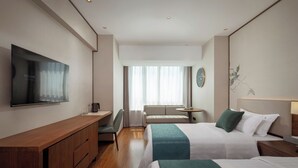 Habitación de hotel con televisión de pantalla plana, escritorio con cajones, sofá, mesa redonda y dos camas con ropa de cama verde y blanca.