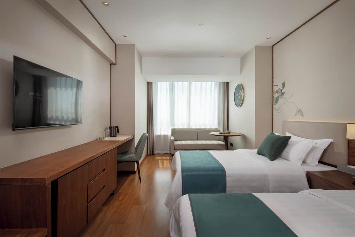 A hotel room with a flat-screen TV, a desk with drawers, a sofa, a round table, and two beds with green and white bedding.