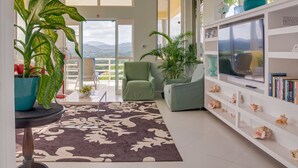 Un salón espacioso con un gran ventanal, un sillón verde, un televisor sobre un estante blanco y una planta en maceta.