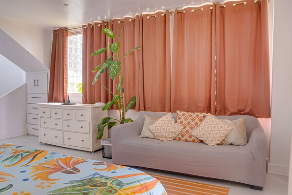 Une chambre à coucher avec un lit, une commode, un canapé, une fenêtre avec des rideaux et une plante.