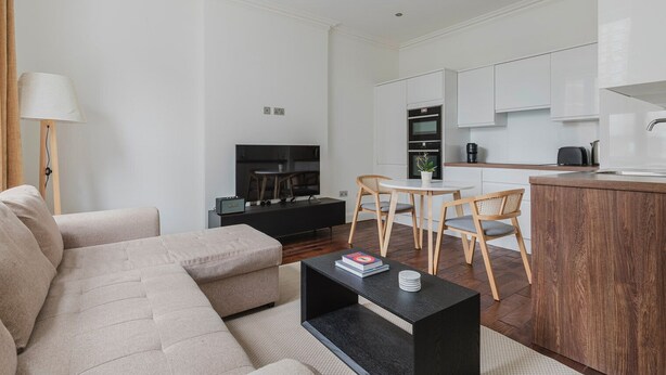 A modern living room with a sofa, a coffee table, and a TV. There is a dining area with a table and chairs. The kitchen is visible with white cabinets and a sink. - Blueground | Bloomsbury 1bd apartment (London)