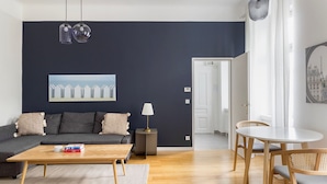 A modern living room with a dark blue accent wall, a sofa with cushions, a coffee table with books, a side table with a lamp, and a dining area with a table and chairs. - Blueground | Mariahilf, fully furnished & ready (Vienna)