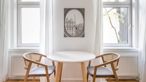A room with a round table and two wicker chairs, a framed picture of a cityscape on the wall, and large windows with white curtains. - Blueground | Mariahilf, fully furnished & ready (Vienna)