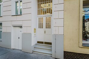 Un bâtiment à la façade blanche, un escalier menant à une porte dotée d’un panneau de verre, et une petite fenêtre donnant sur la rue.