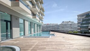 A modern outdoor pool area with a wooden deck, glass balustrades, and a view of adjacent buildings. - Blueground | Landstraße, pool, gym, sauna & a/c (Vienna)