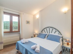 A bedroom with a bed, two bedside lamps, a window with curtains, a radiator, and a small picture on the wall.