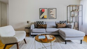 Ein modernes Wohnzimmer mit einer grauen Eckcouch, einem runden Couchtisch und einem weißen Sessel. An der Wand befinden sich abstrakte Gemälde und ein Metallregal mit Dekorationselementen.