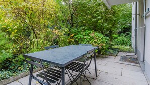 Eine Terrasse mit Tisch und Stühlen, umgeben von üppiger Bepflanzung und einem Gebäude im Hintergrund.