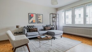 Ein modernes Wohnzimmer mit grauem Sofa, einem weißen Sessel und einem Couchtisch. An der Wand befinden sich zwei abstrakte Gemälde, ein hoher, schwarzer Schrank und ein Fenster mit weißen Vorhängen.