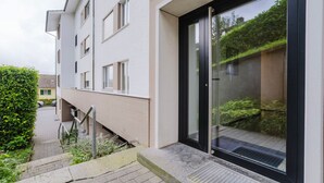 A modern building with a glass door entrance, a visible staircase, and a pathway leading to a green hedge.