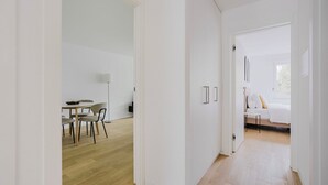 A modern, minimalist room with a wooden floor, white walls, and a bed with white bedding. There is a small table with chairs and a floor lamp in the corner.