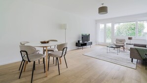 A modern living room with a dining area, a flat-screen TV, and a large window providing natural light.