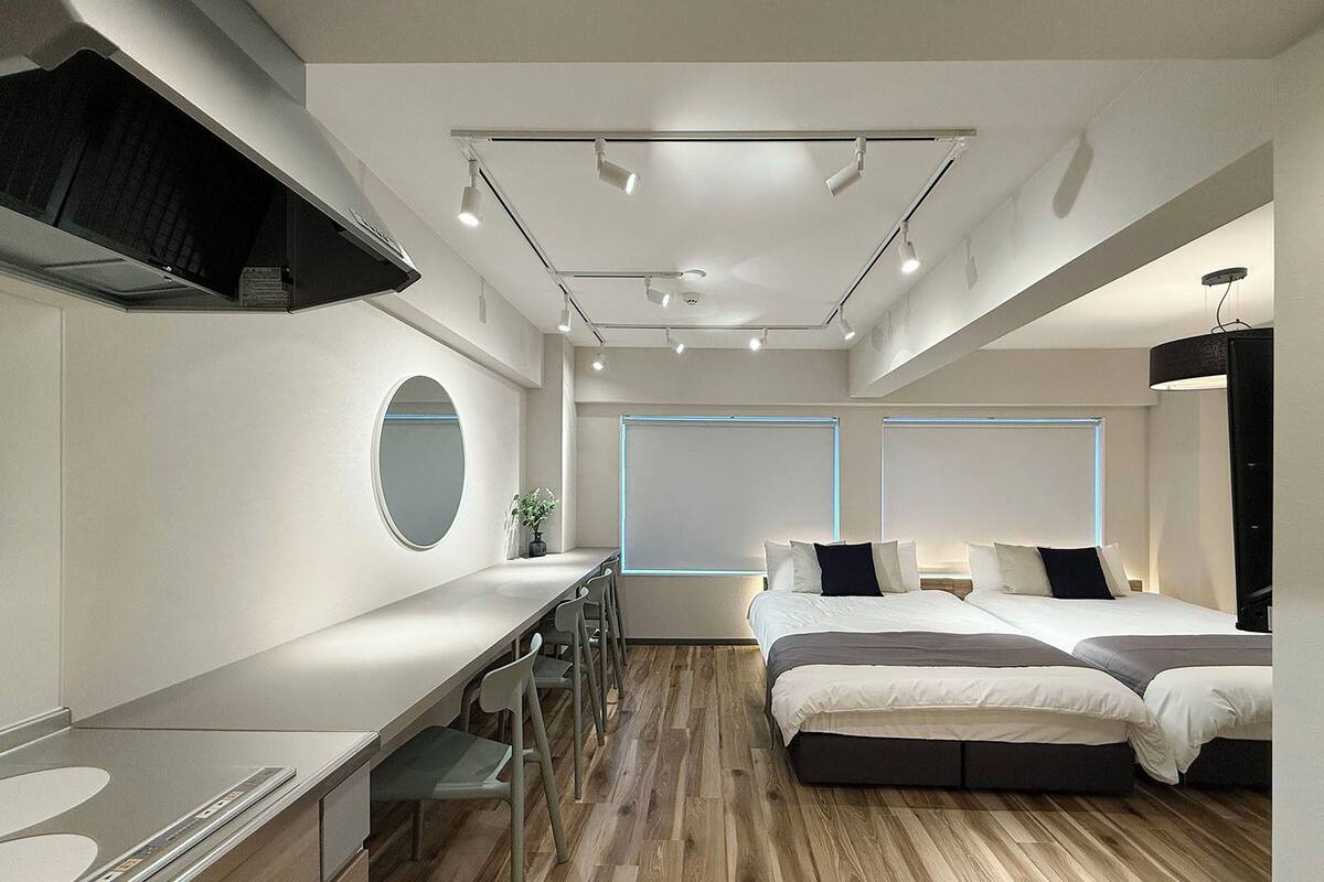 A modern hotel room with a wooden floor, a large bed, a desk with chairs, a round mirror, and a window with blinds.