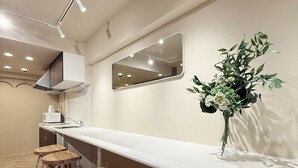 A modern kitchen with a long counter, three wooden stools, a mirror, and a vase with flowers. - Clio Matoba by b-hotel (Hiroshima)
