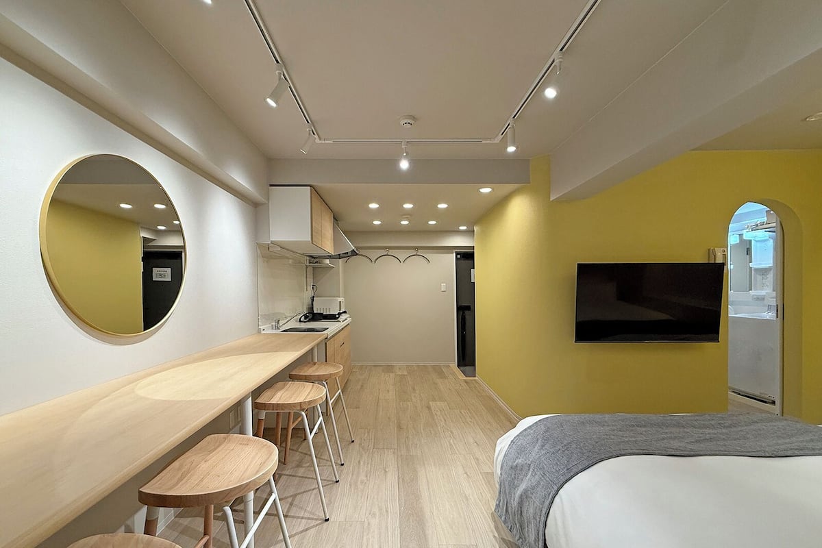 A compact hotel room with a bed, a TV mounted on the wall, a small kitchenette, and a mirror on the wall.
